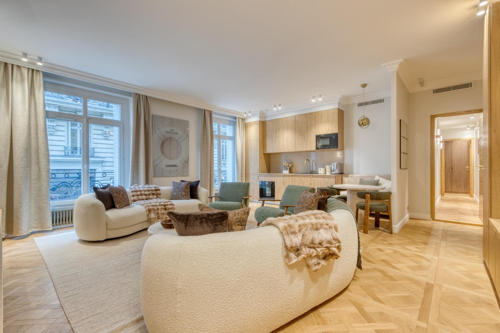 Séjour d'un appartement parisien rénové : parquet en panneaux de Versailles, cuisine en bois clair au fond, fenêtres sur balcon